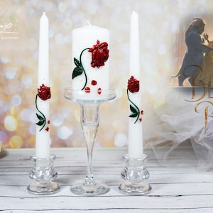 Puede incluir: Tres velas de boda blancas con rosas rojas y hojas verdes. La vela del centro es más alta que las dos velas laterales. Las velas están sobre candelabros de vidrio. El fondo es una imagen borrosa de Bella y la Bestia de la película La Bella y la Bestia.