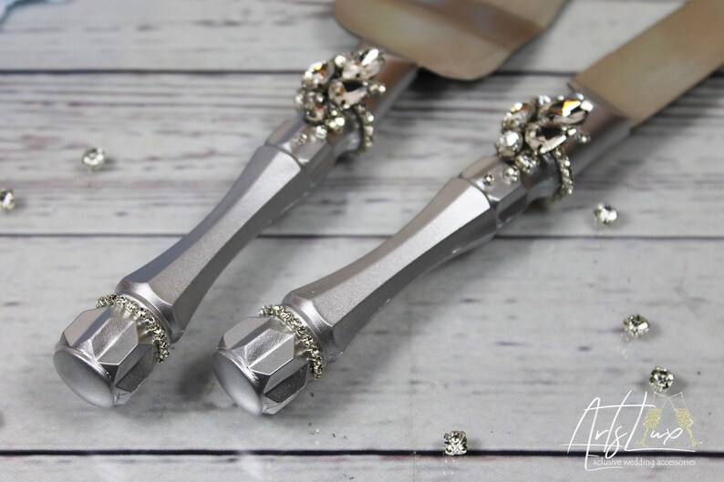 May include: A set of silver wedding cake cutting knives with rhinestone embellishments. The knives have a silver finish and are decorated with sparkling rhinestones.