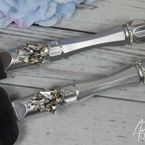 May include: Silver cake knife and server set with crystal embellishments. The handles are silver and have a diamond-shaped pattern. The knife and server are decorated with clear crystals.
