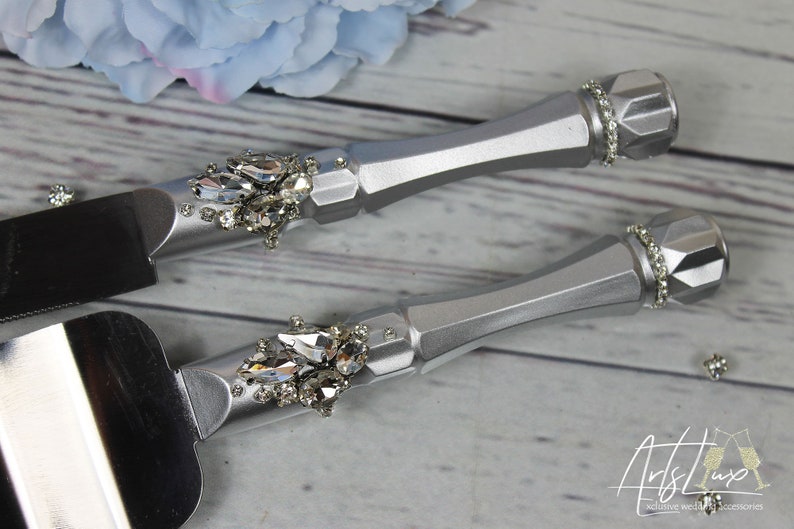 May include: Silver cake knife and server set with crystal embellishments. The handles are silver and have a diamond-shaped pattern. The knife and server are decorated with clear crystals.