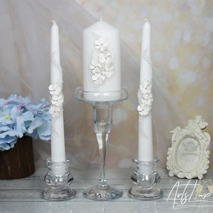Puede incluir: Tres velas de boda blancas con delicados diseños florales. La vela del centro es más alta y tiene un diseño más elaborado. Las velas están sobre candelabros de vidrio transparente.