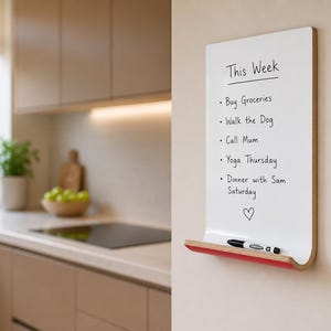 May include: A rectangular white wall-mounted dry erase board with a light wood grain edge and a red shelf at the bottom. The board features the text This Week followed by a list of tasks and a heart symbol. A black marker rests on the red shelf.