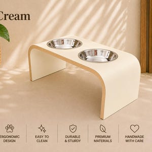 Cream raised dog bowl stand with stainless steel bowls, handmade elevated wooden pet feeder in a bright modern home, elegant feeding station for dogs and cats.