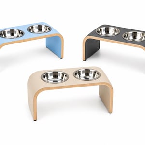 Three handmade double raised pet feeders in Hemp (Beige), Powder Blue, and Graphite Grey finishes, each featuring a modern curved wooden design with two removable stainless steel bowls. Premium elevated feeding stations for cats and dogs with durable, easy-clean laminate surfaces, displayed on a clean white background for easy colour comparison.