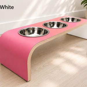 Modern pink and white raised triple dog bowl stand with stainless steel bowls, handcrafted curved wooden pet feeder for dogs and cats.