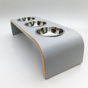 Angled view of our triple bowl raised dog feeder in a light grey finish on a white background with three stainless steel bowls and showing the natural wood edge finish.