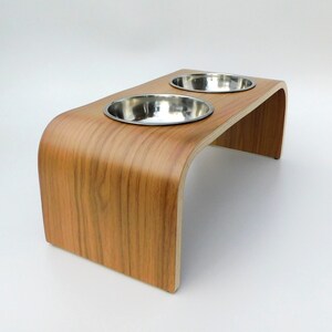 Angled view of a raised dog bowl stand in a walnut finish holding 2 stainless steel bowls and natural wood trim on a white background.