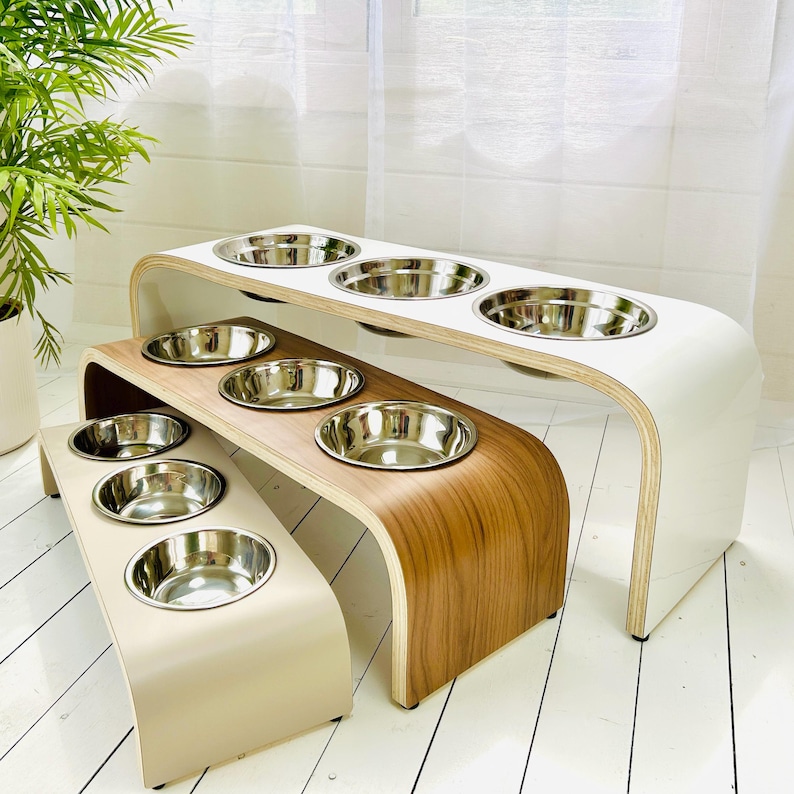 May include: Three tiered pet feeding stations made of bent plywood with smooth, curved edges. Each station features three circular cutouts holding removable stainless steel bowls. The stands are finished in white, natural wood grain, and light beige.