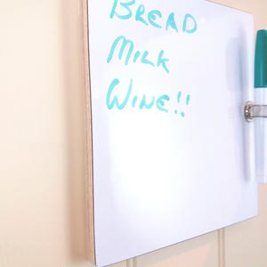 Angled view of small whiteboard / message board / fridge board attached by Velcro pads, pen and pen holder included with a natural wood trim on a white background.