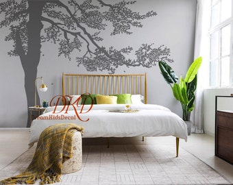May include: A bedroom with a gold metal bed frame and a gray wall with a large tree decal. The tree decal has a gray trunk and branches with green leaves. There is a white blanket on the bed and a green and yellow pillow. There is a small plant in a pot on the floor next to the bed.