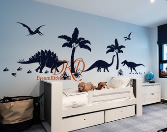 May include: A white twin-sized bed with a white headboard and footboard. The bed has a white mattress and a white bedspread. There are two stuffed animals on the bed. The wall behind the bed is painted a light blue color and has a dinosaur-themed wall decal. The decal features a stegosaurus, a brachiosaurus, a tyrannosaurus rex, and two pterosaurs. The decal also includes palm trees and footprints. The room has a light blue carpet and a wooden toy train set on a table in the foreground.