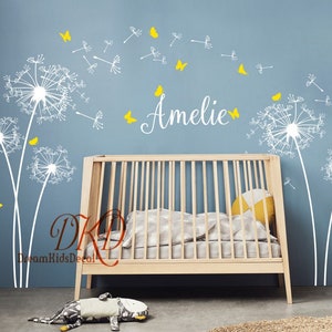 K&ouml;nnte beinhalten: Wei&szlig;e L&ouml;wenzahndeko-Aufkleber mit gelben Schmetterlingen, der Name Amelie in Kursivschrift und ein Holzbabybett im Hintergrund.