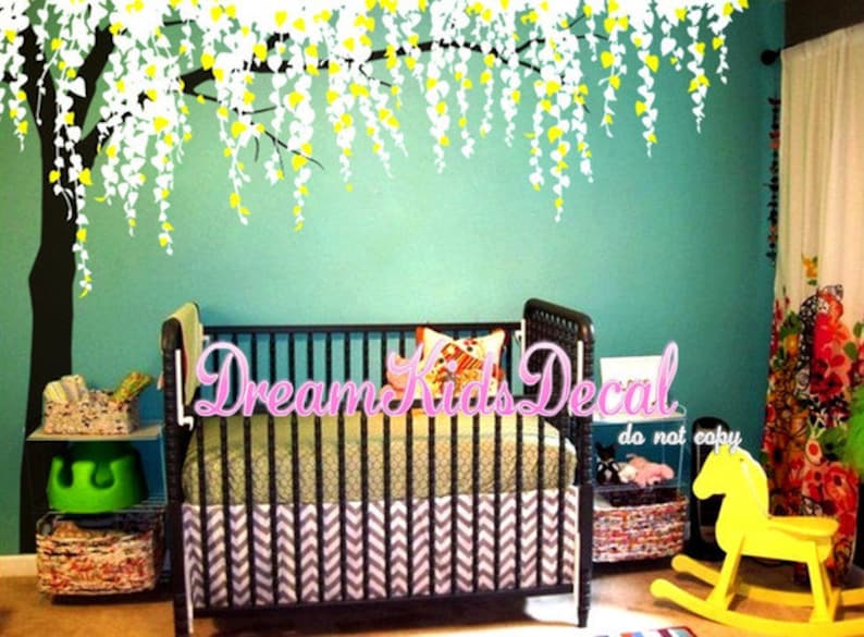 May include: A white and yellow weeping willow tree wall decal above a black crib with a chevron patterned crib skirt. The crib is in a nursery with a green wall and a yellow rocking horse.