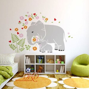 Può includere: Un adesivo da parete con una madre elefante e il suo bambino, in grigio e bianco, con fiori rosa, arancioni e rossi. L'adesivo è su una parete bianca in una cameretta con una poltrona verde, una libreria bianca e un letto verde e bianco.
