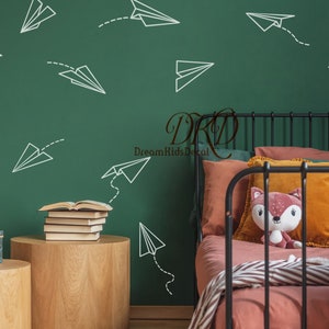 Könnte beinhalten: Eine grüne Wand mit weißen Papierflieger-Aufklebern. Ein schwarzes Metallbettgestell mit einer rosa-weißen gestreiften Decke, einem pfirsichfarbenen Kissen und einem Stofftier-Fuchs. Zwei Holz-Beistelltische mit Bücherstapeln und einer türkisfarbenen Box.