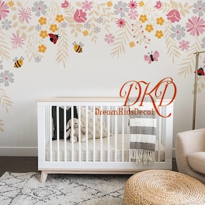 Könnte beinhalten: Ein weißes Babybett mit einer weiß-grauen gestreiften Decke und einem weiß-grauen gemusterten Kissen. Das Bett steht in einem Kinderzimmer mit einer weißen Kommode und einem weißen Teppich. Die Wand ist mit einem floralen Wandtattoo mit Bienen und Marienkäfern verziert.