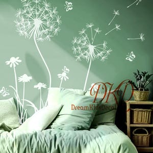 Puede incluir: Calcomanías de pared blancas de diente de león y mariposas sobre una pared verde. Las calcomanías incluyen grandes dientes de león, flores más pequeñas y mariposas. La imagen también muestra una cama con ropa de cama y almohadas verdes, y una pequeña mesita de noche de madera.