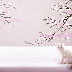 Op de afbeelding: Een witte kat zit op een witte bank voor een muur met een sticker van een roze en witte kersenboom.