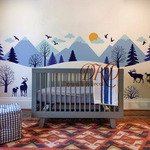 Op de afbeelding: Een kinderkamer met een grijze wieg, een blauw-witte deken en een kleurrijk tapijt. De muur is voorzien van een berg- en bossticker met herten en vogels. Een wereldbol op een houten standaard staat rechts.