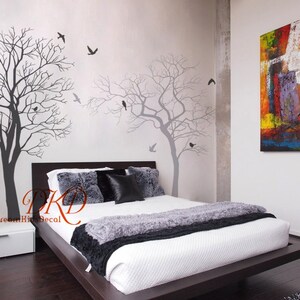 May include: A bedroom featuring a bed, a bedside table, and a large abstract painting on the wall. The walls are adorned with a tree and bird silhouette wall decal. The bed has a white duvet and a black and grey throw blanket.