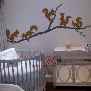 Pode incluir: Um adesivo de parede marrom apresentando um longo galho de árvore com cinco esquilos em várias poses. O adesivo é aplicado em uma parede clara acima de um berço redondo e um trocador. O texto DKD DreamKidsDecal é visível em vermelho.
