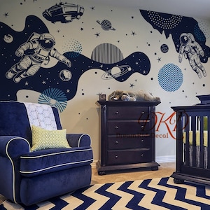 May include: A nursery room with a blue and white chevron rug, a dark wood dresser, a crib, and a blue armchair. The wall is decorated with a space-themed wall decal featuring an astronaut, a spaceship, planets, and stars.