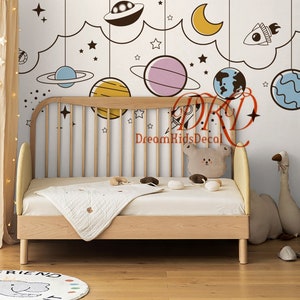 May include: A light wood bed with a white quilt and a bear-shaped cushion. The wall features a space-themed mural with planets, stars, and a rocket. A beige curtain with star lights hangs to the left. The text "DreamKidsDecal" is visible.