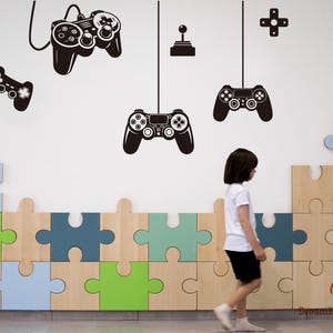 Op de afbeelding: Een muursticker set met zwarte silhouetten van diverse videogamecontrollers, een joystick en een richtingspad. De ontwerpen zijn op een witte muur geplaatst boven een decoratieve lambrisering van grote, in elkaar grijpende houten puzzelstukken.