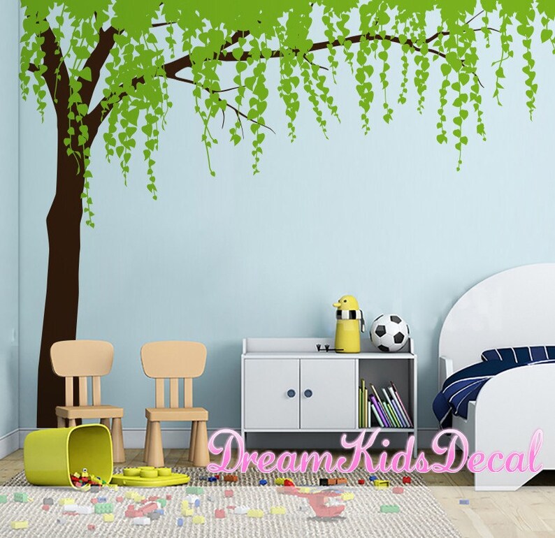 May include: A green tree wall decal with brown branches and leaves. The decal is shown on a light blue wall in a child's bedroom. The room also features a white bed, two wooden chairs, a white cabinet with two doors, and a soccer ball.