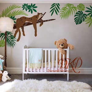 Peut inclure: Décoration murale de chambre d'enfant avec un léopard brun se reposant sur une branche, entouré de feuilles tropicales vertes et vert clair. Un lit bébé blanc avec une couverture bleu clair et un ours en peluche au premier plan.