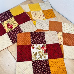 May include: Three square quilted fabric mats featuring a patchwork design of various squares in shades of mustard yellow, deep burgundy, and cream. The patterns include small floral prints, geometric shapes, and a central square with red cherries.