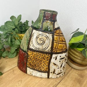 May include: A ceramic vase featuring a patchwork design of various geometric patterns and textures. The surface is divided into sections with colours including green, brown, red, and cream, separated by dark brown lines, creating a mosaic effect.
