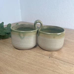 May include: A double-chambered ceramic bowl featuring two connected circular basins with a central arched handle. The exterior and interior surfaces display a speckled cream base colour transitioning into a muted sage green glaze at the top rim.