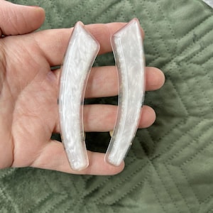 May include: A pair of curved, elongated hair clips featuring a translucent white base with a swirling, pearlescent marbled pattern throughout. The clips have a smooth, glossy finish and a tapered, crescent-like shape with rounded edges.