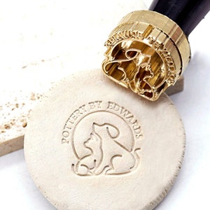 Könnte beinhalten: Ein goldfarbener Stempel aus Metall mit einem schwarzen Griff. Der Stempel zeigt ein Design mit einem Hund und einer Katze, die sich kuscheln. Der Stempel wird verwendet, um einen Abdruck auf einer weißen Tonfläche zu erstellen. Der Abdruck lautet "Pottery by Edwards".
