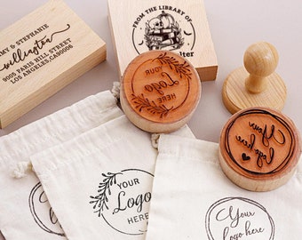 May include: A set of four wooden stamps with different designs. The stamps are round and have a brown wooden handle. The designs include a wreath with the text "Your Logo Here", a design with a book stack and the text "From the Library of", a design with the text "Your Logo Here" and two hearts, and a design with the text "Your Logo Here". The stamps are packaged in white muslin bags.