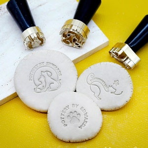 May include: Three clay stamps with black handles. The stamps are for pottery and feature a paw print, a cat, and a dog and cat. The stamps are on a yellow surface.