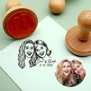 May include: Wooden stamps with a round base and a handle. One stamp has a red ink pad. A stamped image of two smiling women is on a white paper. The text "Dan & Steph 11-12-2025" is below the image. A color photo of the women is also shown.