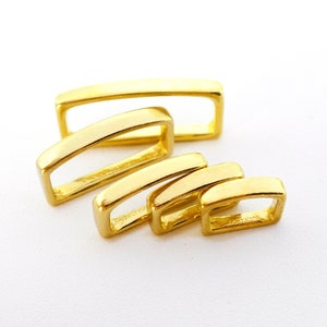 Puede incluir: Conjunto de cuatro trabillas de cinturón de metal dorado de diferentes tamaños. Las trabillas son de forma rectangular y tienen un acabado liso y pulido.