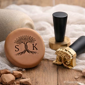 May include: A brown leather stamp with a tree design and the initials "S K". Two black handled stamps with gold and brass accents are also present. The items are on a wooden surface with a white cloth.