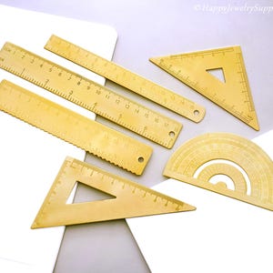 Puede incluir: Un conjunto de instrumentos de dibujo de color latón, que incluyen reglas, triángulos y un transportador. Las reglas están marcadas con medidas en centímetros. Las herramientas están dispuestas sobre una superficie blanca y gris. El texto "HappyJewelrySupplies" es visible.