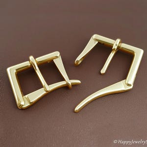 May include: Two shiny gold-colored belt buckles. The buckles are rectangular with a curved bar and a prong. The background is a solid brown color. The buckles are likely for leathercraft or jewelry making.