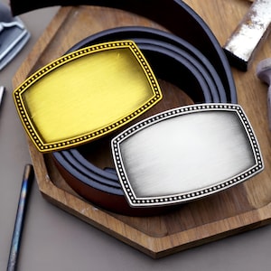 Könnte beinhalten: Zwei rechteckige Gürtelschnallen, eine goldene und eine silberne, mit gepunktetem Rand, liegen auf einem braunen Ledergürtel. Die Schnallen werden auf einem Holztablett präsentiert, was auf Accessoires hindeutet.