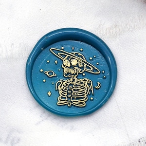 Puede incluir: Un sello de cera turquesa con un diseño de esqueleto dorado que lleva un sombrero con temática espacial. También se ven cuentas de cera turquesa y un mango de sello de cera dorado y rojo. El diseño está sobre una tela blanca.