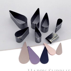 Può includere: Set di otto punzoni in metallo a forma di goccia per cuoio, di diverse dimensioni. I punzoni sono disposti a forma di stella su una superficie grigia. Sotto i punzoni ci sono cinque forme di goccia di cuoio in diversi colori, tra cui beige, viola e blu navy.