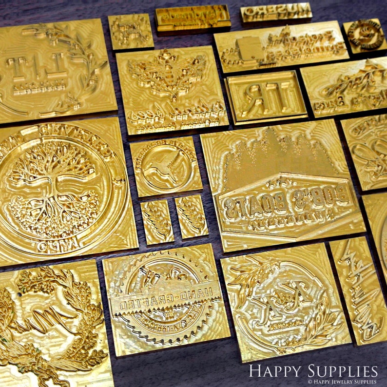 K&ouml;nnte beinhalten: Eine Sammlung von goldfarbenen Metallstempeln mit verschiedenen Designs, darunter ein Baum, ein Vogel, ein handgefertigtes Etikett und ein Boot. Die Stempel sind auf einer Holzoberfl&auml;che angeordnet.