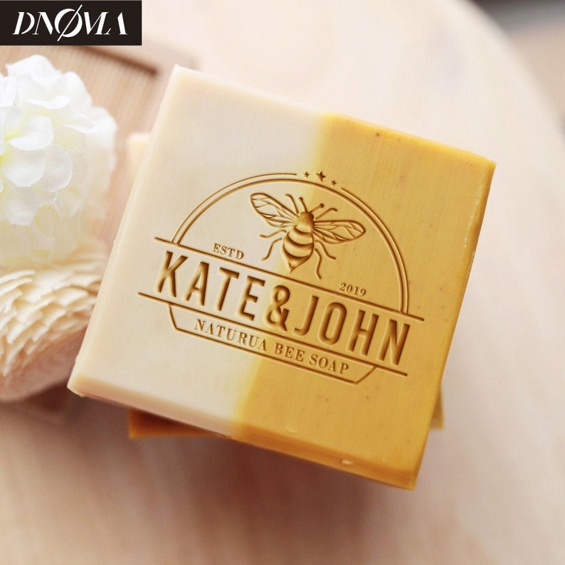 May include: A bar of soap with a yellow and white design. The soap has a logo that says "Kate & John" with a bee and the text "Estd 2019" and "Natura Bee Soap".
