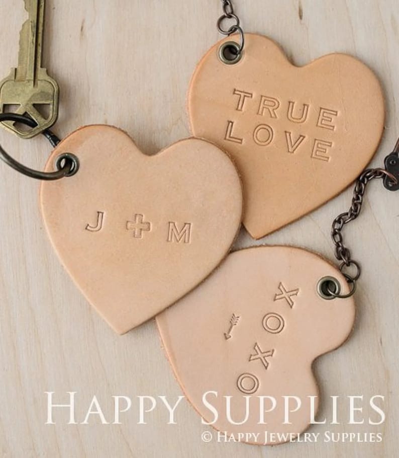 K&ouml;nnte beinhalten: Drei lederne herzf&ouml;rmige Schl&uuml;sselanh&auml;nger mit verschiedenen Gravuren. Ein Schl&uuml;sselanh&auml;nger sagt "True Love", ein anderer sagt "J + M" und der dritte hat einen kleinen Pfeil und "XO" Symbole.