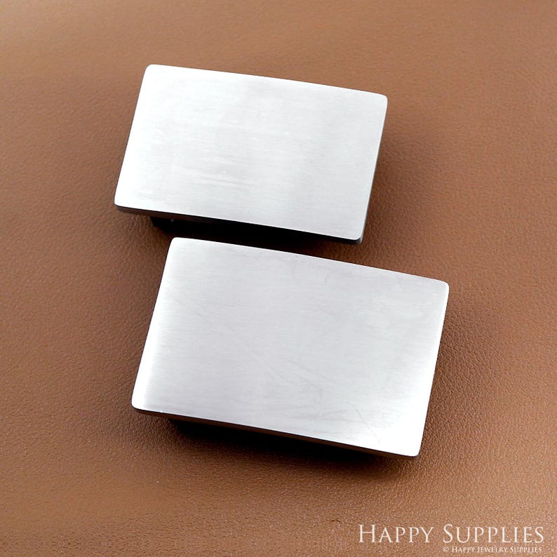 May include: Two rectangular, brushed silver metal belt buckles. The buckles are displayed on a brown leather-like surface. The buckles have a simple, minimalist design.