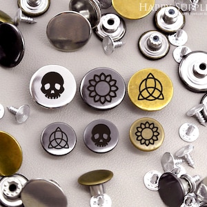 Puede incluir: Surtido de botones a presión de metal plateado y dorado con varios diseños grabados, incluyendo una calavera, un girasol y un símbolo de triquetra. Los botones están dispuestos sobre una superficie blanca. HAPPY SUPPLIES HAPPY JEWELLERY SUPPLIES
